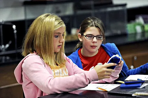 Hands adjust a tablet beside a laptop and worn notebook on a student desk.