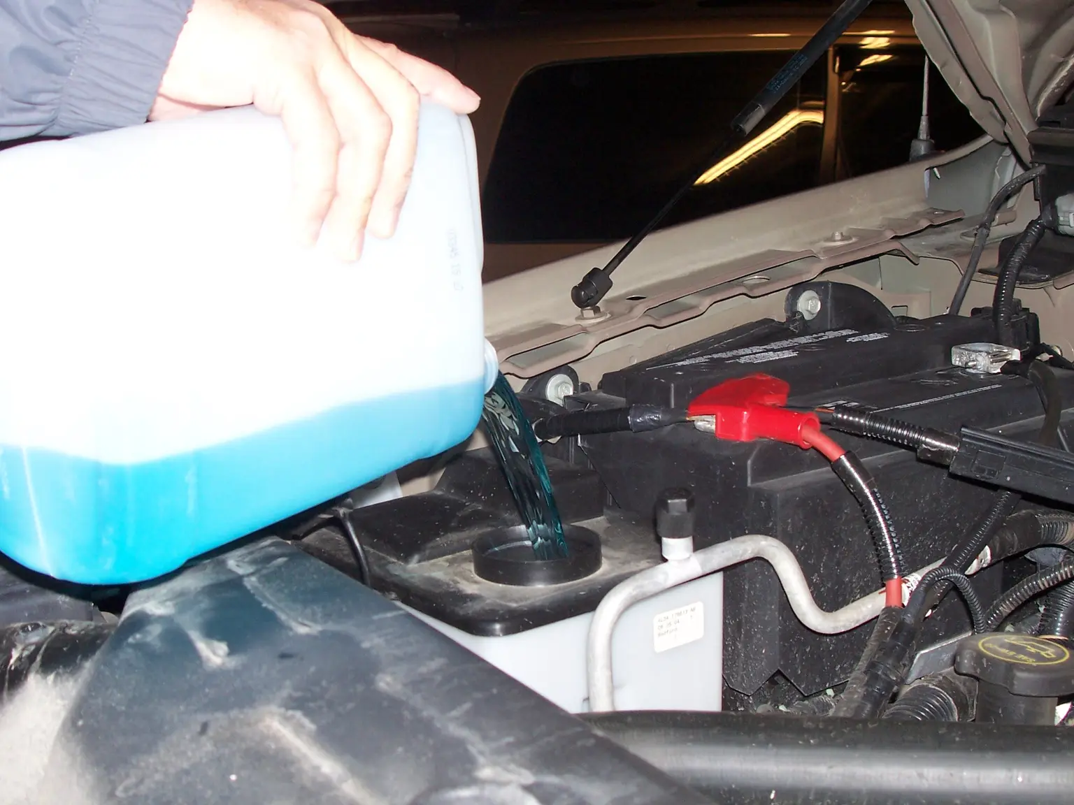 Driver compares two washer fluid jugs beside a car in a snowy driveway with the hood open.